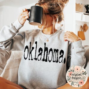 Peut inclure: Un sweat-shirt gris avec le mot "Oklahoma" imprimé en lettres noires en bloc. Le sweat-shirt a un aspect vieilli et délavé. L'image comprend également un petit motif floral avec le texte "original designer."