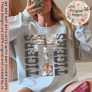May include: A heather gray sweatshirt with the word "TIGERS" in dark gray block letters on either side of a large "T" filled with a camouflage pattern. The text "WE ARE NOT AFFILIATED WITH ANY OTHER SHOPS! These designs are 100% original and created by us." is printed vertically on the left side.