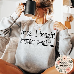 May include: A grey sweatshirt with the text "oops, i bought another t-shirt..." printed on the front. The sweatshirt is being worn by a person holding a black mug.