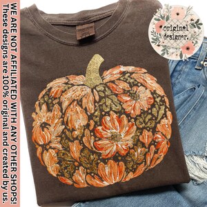 May include: A dark brown t-shirt featuring a large graphic of a pumpkin composed of orange and gold glittery floral patterns. The shirt has a crew neckline and a Comfort Colors brand label at the inner collar. The design includes intricate leaf and petal details in shades of orange, gold, and brown, creating a textured appearance across the center of the garment.