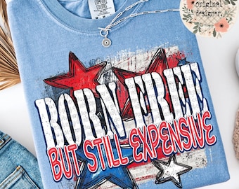 Pode incluir: Camiseta Comfort Colors azul claro com um design patriótico. O gráfico apresenta estrelas vermelhas e azuis e o texto "BORN FREE BUT STILL EXPENSIVE". Um colar de prata com um pingente está drapeado sobre a camiseta.
