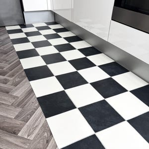May include: A black and white chequered floor in a kitchen. The square tiles create a grid pattern. The floor transitions to a wood-effect herringbone design. White cabinets and a stainless steel appliance are visible.