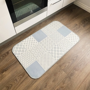 May include: A gray and white patterned kitchen mat with a blue border. The mat is placed in front of a white kitchen cabinet and a stainless steel oven.