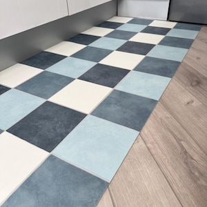 May include: A blue and white chequered floor mat made of square tiles. The mat is placed in front of a white cabinet. The floor next to the mat is made of light brown wood. The mat's design is reminiscent of a chessboard.