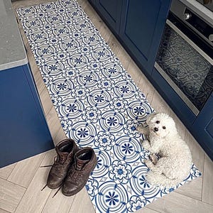 Könnte beinhalten: Ein gemusterter Küchenläufer mit einem sich wiederholenden blau-weißen Blumendesign. Braune Lederstiefel stehen neben dem Teppich auf dem Boden. Ein kleiner weißer Hund liegt auf dem Teppich in der Nähe des Ofens.