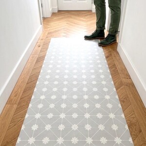Puede incluir: Una alfombra rectangular con fondo gris claro, un patr&oacute;n de cuadr&iacute;cula geom&eacute;trica blanca y peque&ntilde;os detalles florales blancos en cada intersecci&oacute;n. La alfombra est&aacute; colocada a lo largo sobre un suelo de madera con patr&oacute;n de espiga.