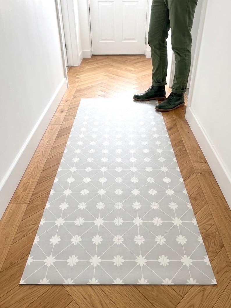 Puede incluir: Una alfombra rectangular con fondo gris claro, un patr&oacute;n de cuadr&iacute;cula geom&eacute;trica blanca y peque&ntilde;os detalles florales blancos en cada intersecci&oacute;n. La alfombra est&aacute; colocada a lo largo sobre un suelo de madera con patr&oacute;n de espiga.