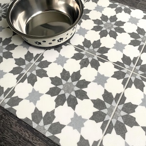 May include: A white and gray patterned pet food mat with a stainless steel dog bowl on top. The mat has a geometric design with black paw prints around the edge of the bowl.