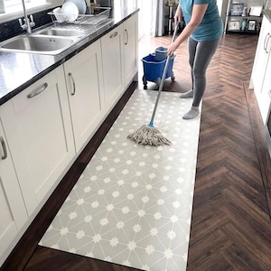 Puede incluir: Una alfombra rectangular con fondo gris claro, un patr&oacute;n de rejilla geom&eacute;trica blanca y peque&ntilde;os detalles florales blancos en cada intersecci&oacute;n, colocada sobre un suelo de madera marr&oacute;n oscuro en espiga en una cocina.