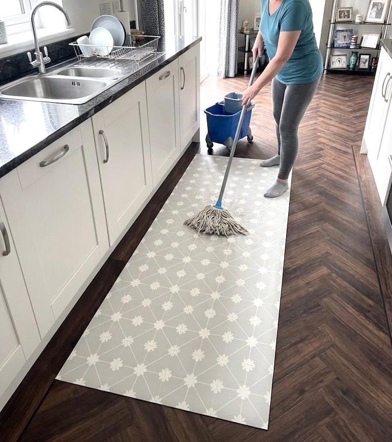 Puede incluir: Una alfombra rectangular con fondo gris claro, un patr&oacute;n de rejilla geom&eacute;trica blanca y peque&ntilde;os detalles florales blancos en cada intersecci&oacute;n, colocada sobre un suelo de madera marr&oacute;n oscuro en espiga en una cocina.