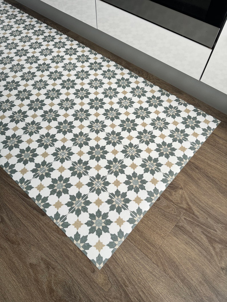 May include: A white, gray, and beige patterned kitchen rug with a geometric design. The rug is placed on a wood floor in front of a white cabinet.