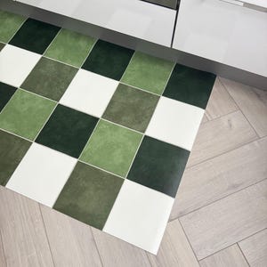May include: A green, white, and dark green checkered kitchen mat. The mat is made of a durable material and is placed on a light wood floor. The mat is near a white cabinet and a stainless steel appliance.