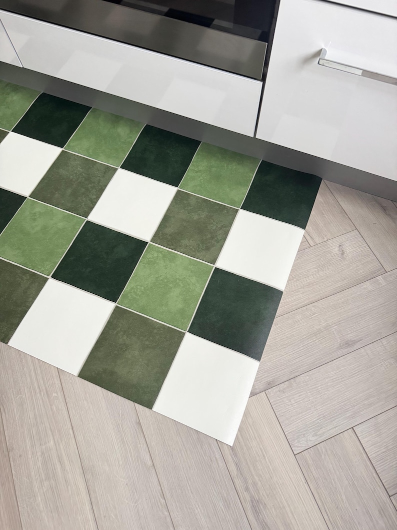 May include: A green, white, and dark green checkered kitchen mat. The mat is made of a durable material and is placed on a light wood floor. The mat is near a white cabinet and a stainless steel appliance.
