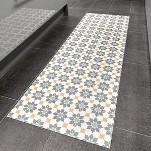 May include: A rectangular kitchen runner rug with a repeating floral pattern in white, blue, and tan. The rug is placed on a dark gray tiled floor, next to a gray cabinet.
