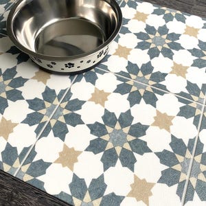 May include: A white and blue patterned pet food mat with a stainless steel dog bowl with black paw prints on the rim. The bowl is sitting on the mat.