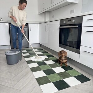 May include: A woman mops a kitchen floor with a green, white, and black checkered rug. A brown dachshund dog sits on the rug near the oven. The kitchen has white cabinets and appliances.