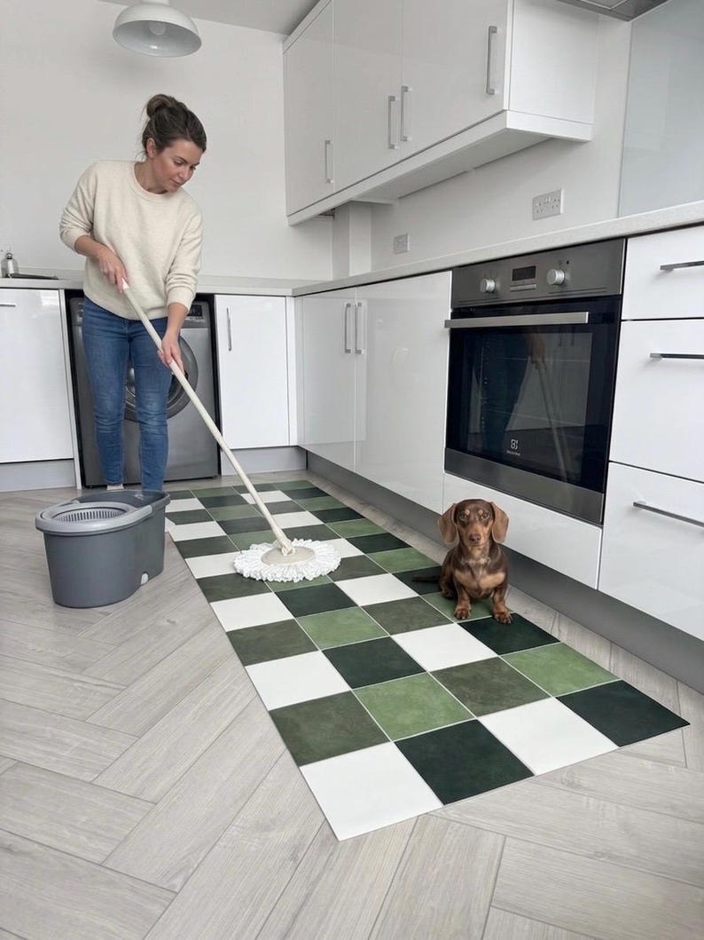 May include: A woman mops a kitchen floor with a green, white, and black checkered rug. A brown dachshund dog sits on the rug near the oven. The kitchen has white cabinets and appliances.