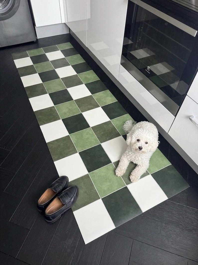 May include: A rectangular kitchen mat with a checkerboard pattern in shades of green and white. Black leather loafers are placed near the mat. A small white dog is lying on the mat.
