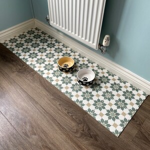 May include: Two dog bowls, one yellow and one white, with black paw prints on a green and beige patterned floor mat. The mat is on a wood floor.