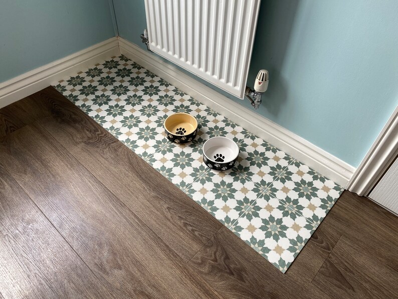 May include: Two dog bowls, one yellow and one white, with black paw prints on a green and beige patterned floor mat. The mat is on a wood floor.