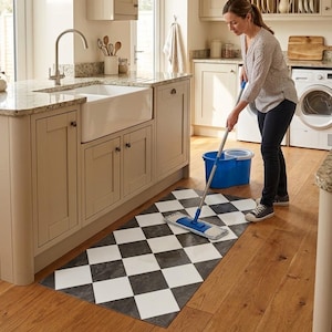 May include: A rectangular floor mat featuring a black and white checkered pattern. The mat is positioned on a wooden floor in a kitchen setting, with a blue mop and bucket nearby. The design consists of alternating dark and light diamond shapes.