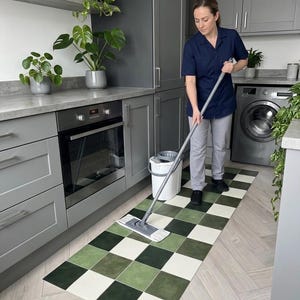 May include: A person mops a kitchen floor with a green, white, and dark green checkered mat. The kitchen has gray cabinets, a stainless steel oven, and a washing machine. Potted plants are on the countertop and shelves.
