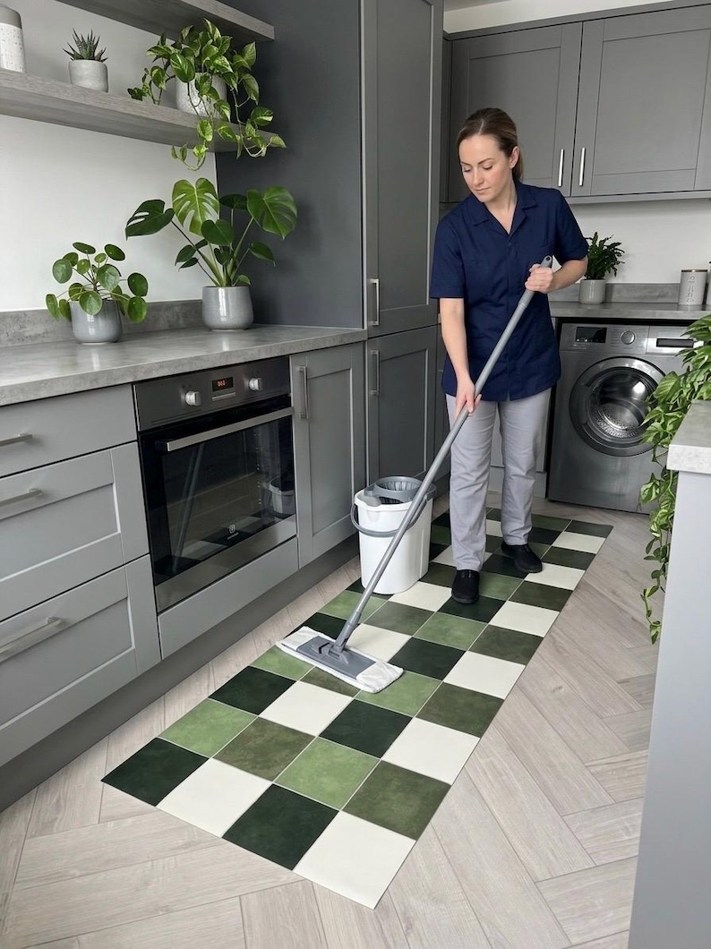 May include: A person mops a kitchen floor with a green, white, and dark green checkered mat. The kitchen has gray cabinets, a stainless steel oven, and a washing machine. Potted plants are on the countertop and shelves.