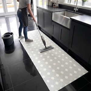 Puede incluir: Una alfombra de suelo larga y rectangular con fondo gris claro y un patr&oacute;n de cuadr&iacute;cula geom&eacute;trica blanca repetitiva con peque&ntilde;os detalles florales. La alfombra est&aacute; colocada sobre un suelo de baldosas oscuras y brillantes.
