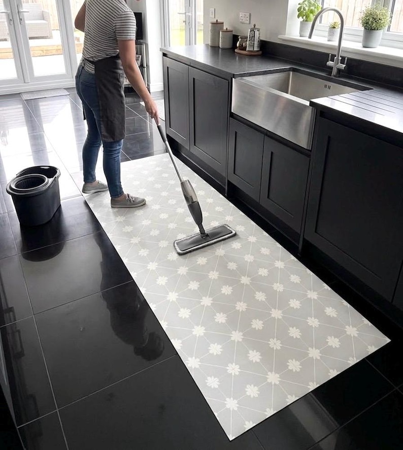 Puede incluir: Una alfombra de suelo larga y rectangular con fondo gris claro y un patr&oacute;n de cuadr&iacute;cula geom&eacute;trica blanca repetitiva con peque&ntilde;os detalles florales. La alfombra est&aacute; colocada sobre un suelo de baldosas oscuras y brillantes.