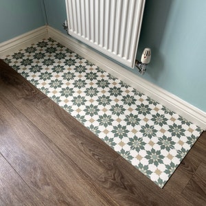 May include: A close-up of a bathroom floor with a patterned tile runner. The runner is made of white, green, and beige tiles with a floral design. The floor is made of dark brown wood laminate.