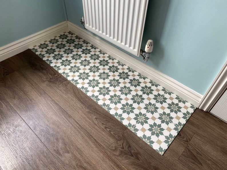 May include: A close-up of a bathroom floor with a patterned tile runner. The runner is made of white, green, and beige tiles with a floral design. The floor is made of dark brown wood laminate.