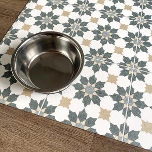 May include: A stainless steel pet food bowl on a white and gray patterned floor mat. The mat has a geometric design with a repeating star pattern.
