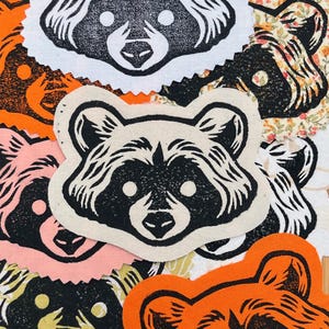 May include: A collection of raccoon-shaped fabric patches in various colors, including white, orange, pink, and cream. Each patch features a black linocut-style raccoon face with detailed fur and facial features. The patches are arranged on a wooden surface.
