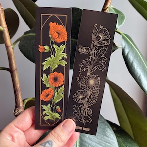 May include: Two black bookmarks with floral designs. One features orange poppies with green leaves within a gold frame. The other has a gold outline of poppies. The text "SPORT STUDIO" is printed on the second bookmark.