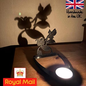 May include: A black metal fairy and flower silhouette tea light holder. The design casts a shadow of a fairy and flowers on the wall. The text "Handmade in the UK" is visible in the upper right corner.