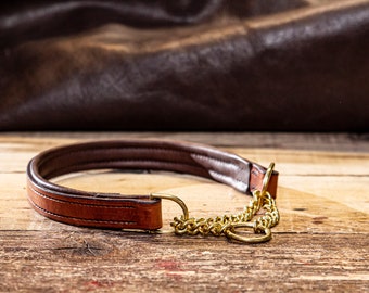 May include: A brown leather dog collar with a brass chain and hardware. The collar is padded and stitched, resting on a wooden surface. The background features a leather texture.