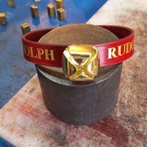 Puede incluir: Un brazalete de cuero rojo con una campana dorada y las palabras "OLPH" y "RUD" en letras doradas.