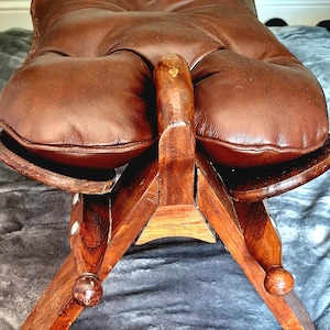 Puede incluir: Un taburete de cuero marrón estilo silla de montar con un cojín de asiento acolchado y capitoné, apoyado sobre una estructura de madera oscura con cuatro patas inclinadas y remates redondeados en la base, con un elemento central de madera.