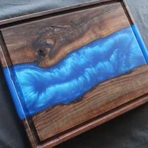 May include: A wooden cutting board with a blue epoxy resin inlay. The board has a raised edge and a groove around the perimeter.