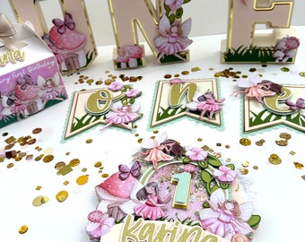 May include: A pink and green fairy themed first birthday cake topper with a large number one, a banner with the letter "o" and "n" and the name "Karina" in gold glitter letters.
