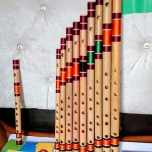 May include: A collection of bamboo flutes of varying lengths, each adorned with burgundy, orange, and green decorative bands. The flutes are arranged in a staggered row, with one flute standing alone. A book titled "Enrich Your Grammar" is visible.
