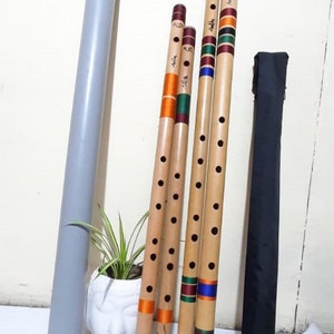 Può includere: Quattro flauti in bambù con fori per le dita e fasce decorative colorate in arancione, verde, blu e bordeaux. I flauti sono posizionati accanto a un tubo cilindrico grigio, un vaso in ceramica bianca a forma di testa con una pianta e una borsa nera.