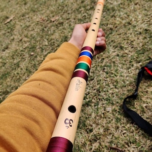 May include: A brown wooden flute with a red, green, blue and orange striped band. The flute is decorated with a maroon ribbon and the word "Halim" is written on the side. The flute is held in a person's hand.