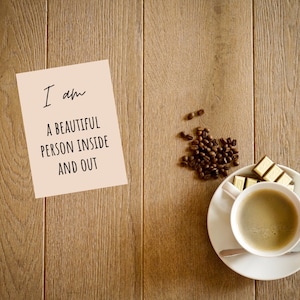 Op de afbeelding: Een beige kaart met de tekst "Ik ben een mooi persoon van binnen en buiten" op een houten oppervlak met een kop koffie en chocoladevierkanten.