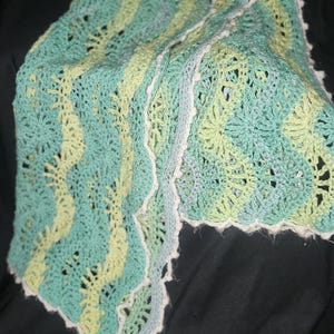May include: A crocheted blanket featuring a wavy, ripple pattern in alternating stripes of light green, teal, and soft blue. The edges are finished with a delicate white scalloped border. The text StitchyWitchByKelly is printed in the corner.