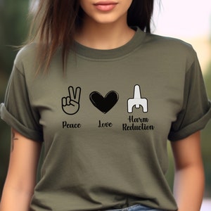 May include: Olive green t-shirt featuring the words "Peace", "Love", and "Harm Reduction" with corresponding black and white graphics. The shirt has short sleeves and a crew neck.