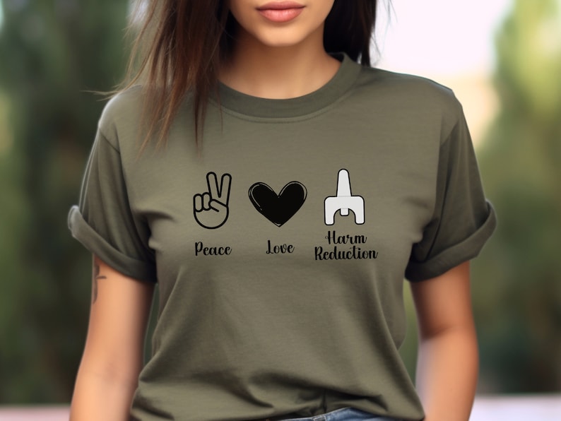 May include: Olive green t-shirt featuring the words "Peace", "Love", and "Harm Reduction" with corresponding black and white graphics. The shirt has short sleeves and a crew neck.