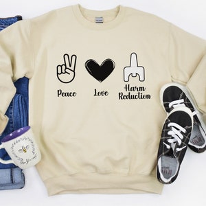 May include: A cream-colored sweatshirt with the words "Peace Love Harm Reduction" and corresponding symbols. The sweatshirt is paired with blue jeans, black and white sneakers, and a mug with a bee design.