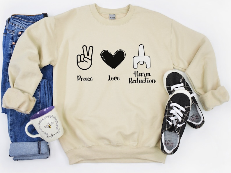 May include: A cream-colored sweatshirt with the words "Peace Love Harm Reduction" and corresponding symbols. The sweatshirt is paired with blue jeans, black and white sneakers, and a mug with a bee design.