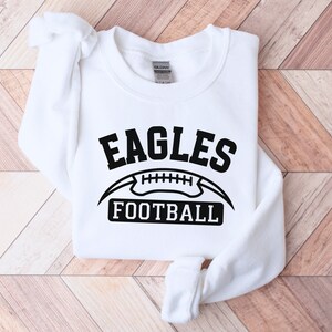 May include: White sweatshirt with black text that reads "Eagles Football" and a black football graphic.